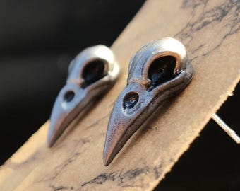 May include: A pair of silver crow skull earrings. The earrings are small and delicate, and they have a realistic design.