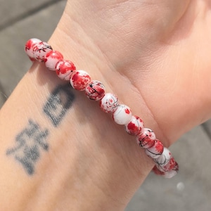 May include: A stretch bracelet composed of round beads featuring a white base with irregular red splatters and fine black vein-like markings. The beads are arranged in a single strand, creating a continuous circular pattern around the wrist.