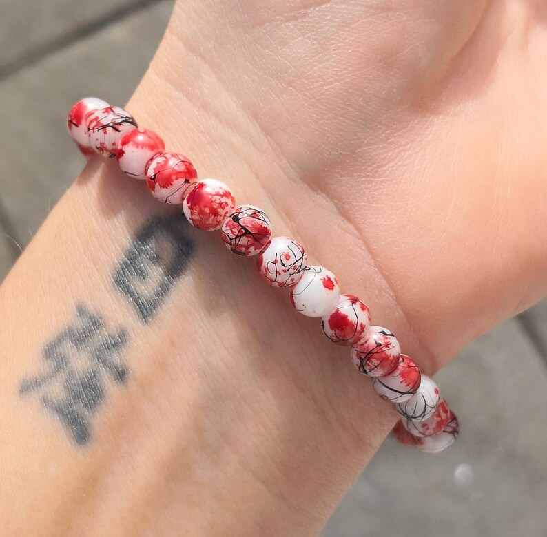 May include: A stretch bracelet composed of round beads featuring a white base with irregular red splatters and fine black vein-like markings. The beads are arranged in a single strand, creating a continuous circular pattern around the wrist.