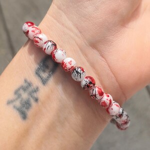 May include: A beaded bracelet featuring spherical white beads with a distinct pattern of red splatters and thin black lines resembling veins or cracks. The beads are strung together in a single row, creating a continuous circular design around the wrist.