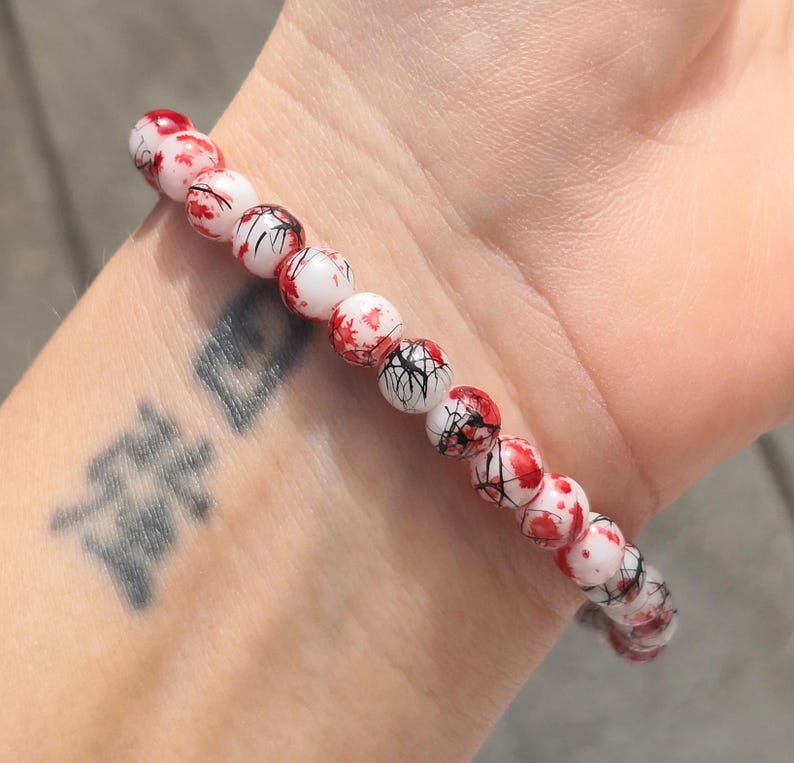 May include: A beaded bracelet featuring spherical white beads with a distinct pattern of red splatters and thin black lines resembling veins or cracks. The beads are strung together in a single row, creating a continuous circular design around the wrist.