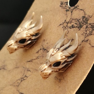 May include: A pair of dragon skull earrings made of white resin with black eyes. The earrings are displayed on a brown background.