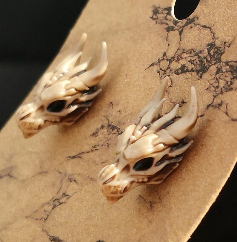 May include: A pair of dragon skull earrings made of white resin with black eyes. The earrings are displayed on a brown background.