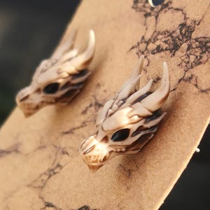 May include: A pair of dragon head stud earrings. The earrings are made from a cream-coloured material and have a detailed design. The dragon heads have black eyes and a textured surface.