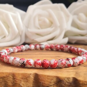 May include: A beaded bracelet featuring small, round, white beads with a distinct red splatter pattern and fine black veining throughout. The beads are strung together in a continuous circular loop, creating a uniform and repetitive design.