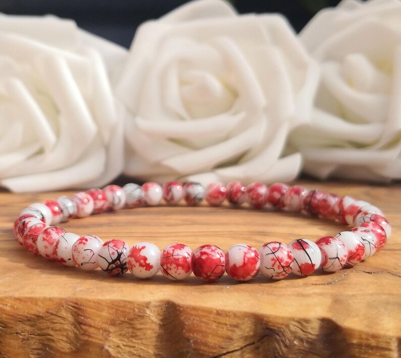 May include: A beaded bracelet featuring small, round, white beads with a distinct red splatter pattern and fine black veining throughout. The beads are strung together in a continuous circular loop, creating a uniform and repetitive design.