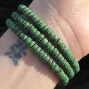 May include: Three green beaded bracelets. Each bracelet is made of small, round, green beads with a slightly weathered appearance. The bracelets are stacked on top of each other, creating a layered effect. The beads have a mottled, dark green and brown colour.