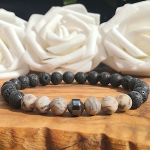 May include: A beaded bracelet featuring a series of round, porous black lava stone beads, interspersed with smooth, light beige patterned stone beads and a single faceted, dark metallic-toned bead positioned at the centre of the strand.
