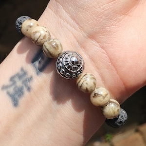 May include: A bracelet with brown and white beads and a silver metal bead with a decorative design. The bracelet is on a person's wrist.