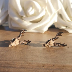 May include: Two small, detailed deer skull earrings with antlers. The earrings are made of a light brown resin and have a realistic look. The earrings are on a wooden surface.