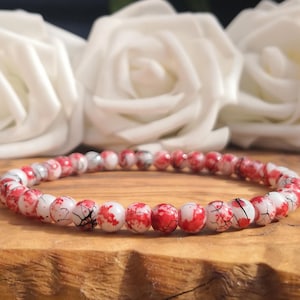 May include: A beaded bracelet featuring small, round, polished beads with a white base color and irregular red splatter patterns, accented by thin black crackle lines throughout the surface of each individual bead in the circular arrangement.