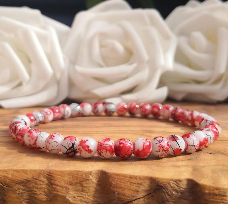 May include: A beaded bracelet featuring small, round, polished beads with a white base color and irregular red splatter patterns, accented by thin black crackle lines throughout the surface of each individual bead in the circular arrangement.