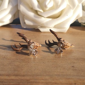 May include: A pair of brown deer head stud earrings with silver posts. The deer heads are detailed with antlers and a textured finish.