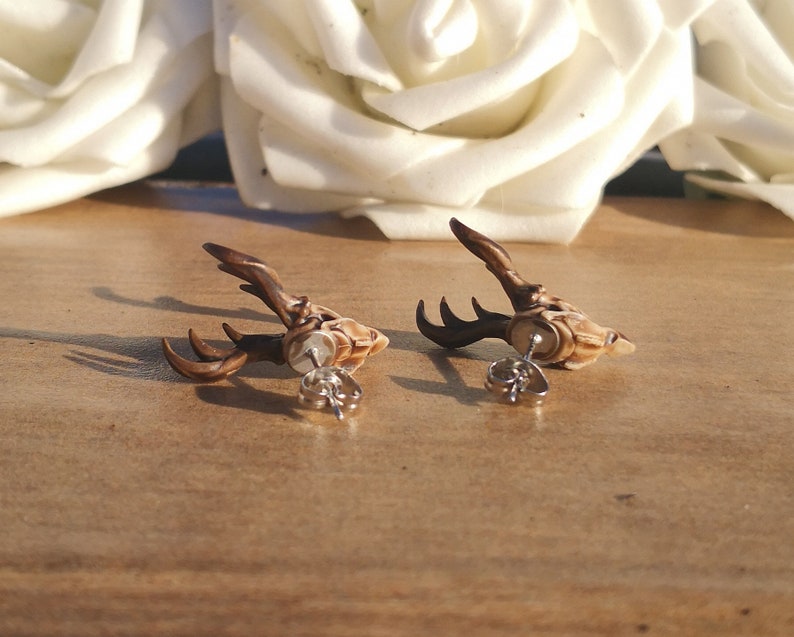 May include: A pair of brown deer head stud earrings with silver posts. The deer heads are detailed with antlers and a textured finish.
