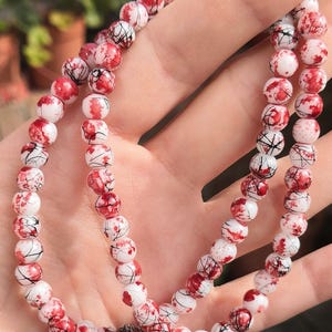 May include: A beaded necklace or bracelet featuring round white beads with a distinct red splatter pattern and thin black crackle lines throughout. The beads are uniform in size and strung together in a continuous loop, creating a high-contrast design.