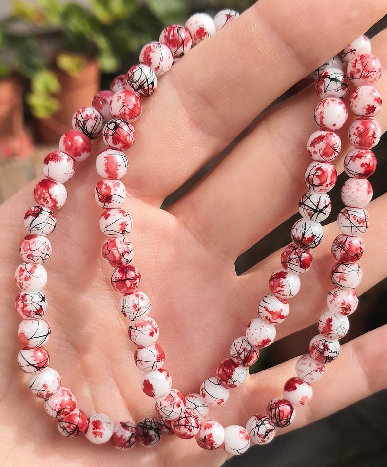 May include: A beaded necklace or bracelet featuring round white beads with a distinct red splatter pattern and thin black crackle lines throughout. The beads are uniform in size and strung together in a continuous loop, creating a high-contrast design.