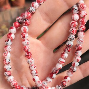 May include: A beaded bracelet featuring round white beads with a speckled red and black pattern. The beads are arranged in a continuous circular strand, showcasing a smooth, polished finish with intricate, thin black lines and vibrant red splotches.