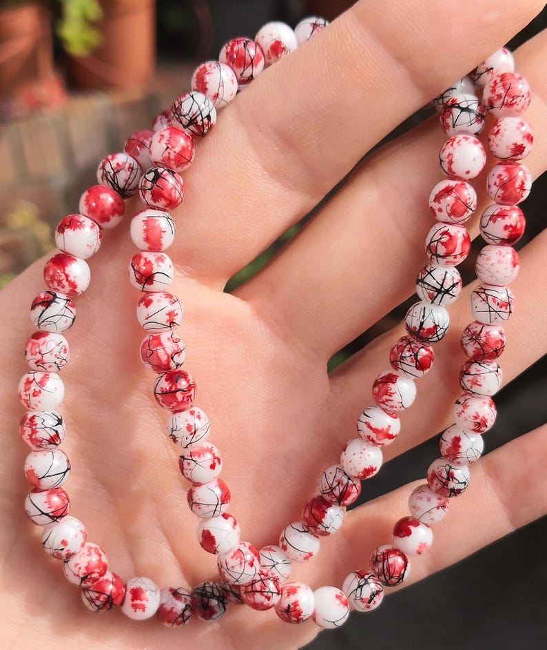 May include: A beaded bracelet featuring round white beads with a speckled red and black pattern. The beads are arranged in a continuous circular strand, showcasing a smooth, polished finish with intricate, thin black lines and vibrant red splotches.