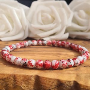 May include: A beaded bracelet featuring small, round, white beads with a vibrant red and black marbled or crackled pattern. The beads are strung together in a continuous circular loop, creating a uniform design that displays a high-contrast color scheme.