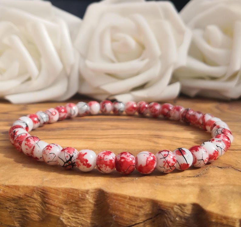 May include: A beaded bracelet featuring small, round, white beads with a vibrant red and black marbled or crackled pattern. The beads are strung together in a continuous circular loop, creating a uniform design that displays a high-contrast color scheme.