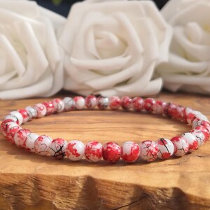 May include: A beaded bracelet featuring small, round, white beads with a vibrant red splatter pattern and fine black crackle detailing throughout. The beads are strung together in a continuous circular loop, creating a uniform and detailed design.