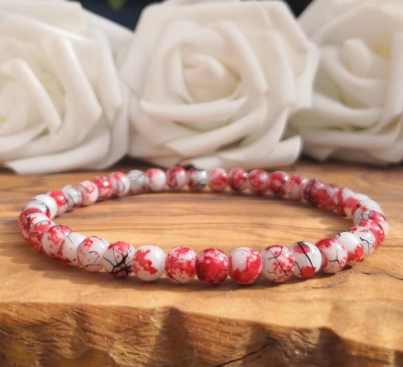 May include: A beaded bracelet featuring small, round, white beads with a vibrant red splatter pattern and fine black crackle detailing throughout. The beads are strung together in a continuous circular loop, creating a uniform and detailed design.