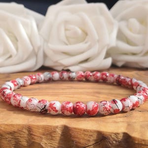 May include: A circular beaded bracelet composed of small, round, white beads featuring a distinct red splatter pattern and fine black crackle lines throughout. The beads are strung together in a single continuous loop, creating a uniform design.
