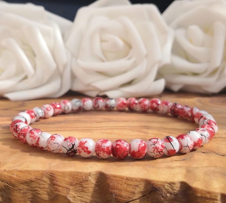 May include: A circular beaded bracelet composed of small, round, white beads featuring a distinct red splatter pattern and fine black crackle lines throughout. The beads are strung together in a single continuous loop, creating a uniform design.