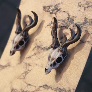 May include: A pair of deer skull earrings with brown antlers. The skulls are white with black eye sockets.