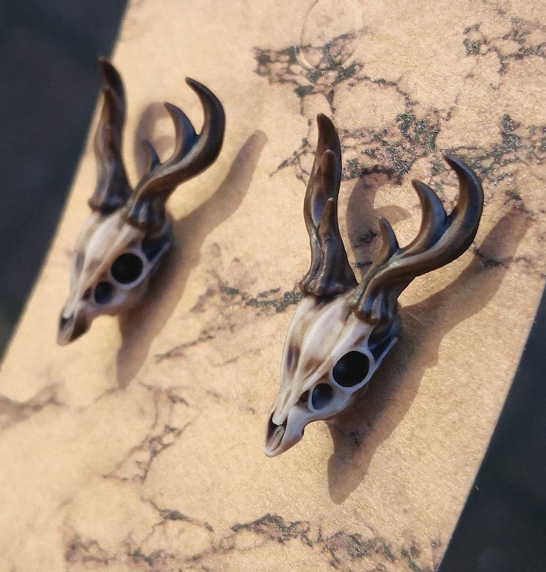 May include: A pair of deer skull earrings with brown antlers. The skulls are white with black eye sockets.