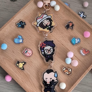 Könnte beinhalten: Ein Set aus drei transparenten Acryl-Schlüsselanhängern mit Cartoon-Charakteren, verbunden durch einen roségoldenen Verschluss. Die Schlüsselanhänger liegen auf einem Holztablett mit kleinen, bunten Pompons und Charakter-Stickern.