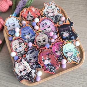 May include: A collection of 12 colourful anime character keychains with different designs. The keychains are made of clear acrylic and feature various characters with different hairstyles, clothing, and accessories. The keychains are displayed on a wooden tray with white pom-poms.
