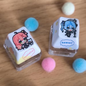 Könnte beinhalten: Zwei weiße Tastenkappen mit Illustrationen und Text im Anime-Stil. Eine zeigt einen pinkhaarigen Charakter mit einer Katze und dem Text "nyahello!". Die andere hat einen blauhaarigen Charakter und das Wort "kawaii!". Mehrere bunte Pompons umgeben die Tastenkappen.