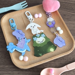 May include: Three acrylic keychains featuring cartoon characters. One is blue with a cloud and creature, one is green with a floral-crowned creature and white rabbit, and one is purple with a heart, a small creature, and a cloud, all with small pom-poms.