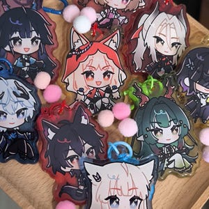 May include: A collection of anime-style character keychains with various designs, including characters with animal ears. The keychains are colorful, with pink, blue, and green accents. Each keychain has a metal ring and a pom-pom attached.