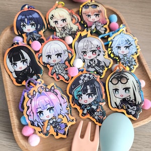 May include: A set of ten colorful anime character keychains with holographic accents. The keychains are arranged on a wooden tray with a fork and spoon.