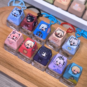 May include: A collection of custom keycaps with anime-style character designs. Each keycap features a unique character illustration in various colors, mounted on a clear, rectangular base. Keycaps are displayed on a wooden tray.