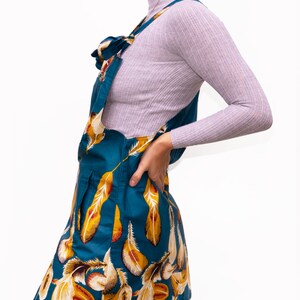 May include: Teal overall dress with a feather pattern in yellow and white. The dress has a bib front and adjustable straps. The model is wearing a light purple long-sleeve shirt underneath. The dress is a mini length.