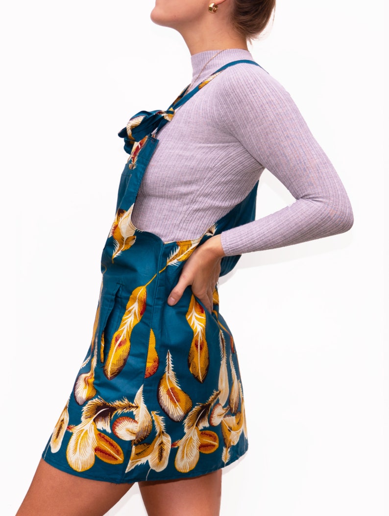 May include: Teal overall dress with a feather pattern in yellow and white. The dress has a bib front and adjustable straps. The model is wearing a light purple long-sleeve shirt underneath. The dress is a mini length.