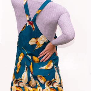 May include: Teal-colored overall dress with a feather pattern in yellow, white, and red. The dress has a bib front, two pockets, and adjustable straps. The model is wearing a gray long-sleeve shirt underneath.
