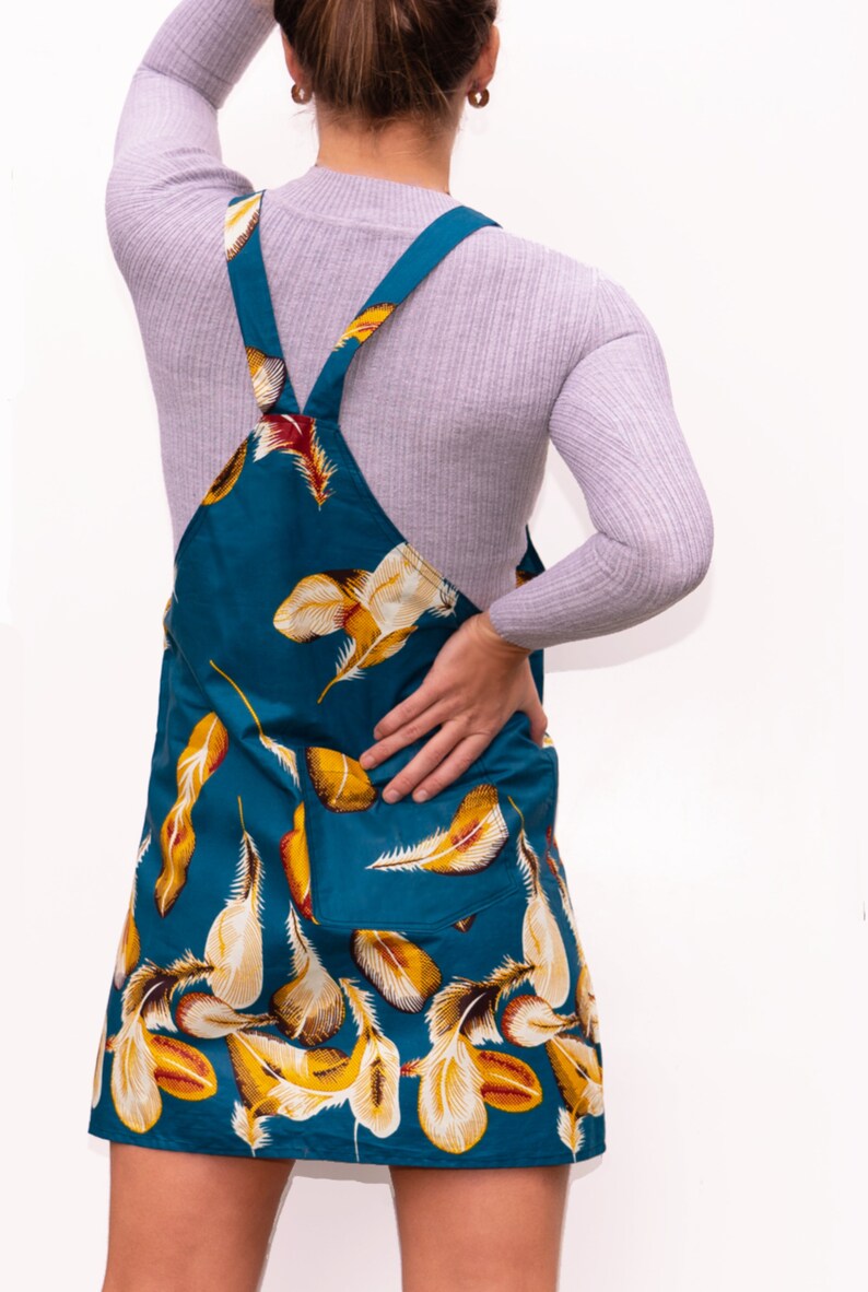 May include: Teal-colored overall dress with a feather pattern in yellow, white, and red. The dress has a bib front, two pockets, and adjustable straps. The model is wearing a gray long-sleeve shirt underneath.