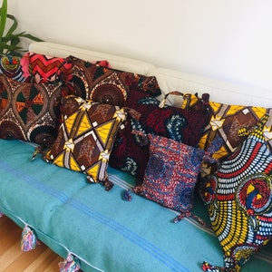 Op de afbeelding: Een verzameling vierkante sierkussens met levendige Afrikaanse waxprint stoffen hoezen. De kussens hebben verschillende patronen in rood, geel, bruin en blauw. Ze zijn gerangschikt op een turquoise kussen met kwastjes, wat een kleurrijk en uitnodigend geheel creëert.