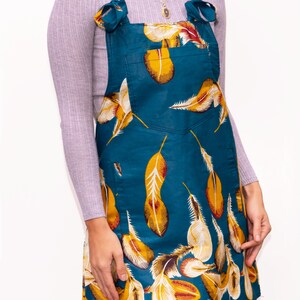 May include: Teal overall dress with a feather pattern in yellow, white, and red. The dress has a front pocket and tie straps. The model is wearing a light purple turtleneck underneath.