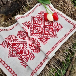 Puede incluir: Un camino de mesa de lino blanco con un diseño bordado en rojo de flores estilizadas y patrones geométricos. El camino de mesa está doblado por la mitad y tiene un borde blanco.