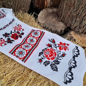 Puede incluir: Tela blanca con diseños florales bordados en rojo y negro, incluyendo rosas y uvas. La tela está sobre un lecho de paja.