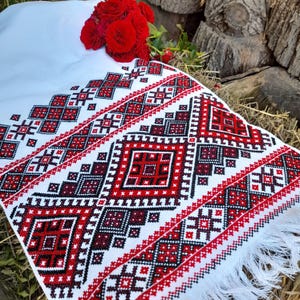 May include: White fabric with red and black embroidered geometric patterns. The fabric has a fringe border.