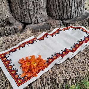 Könnte beinhalten: Ein weißer Tischläufer aus Leinen mit einem roten, orangefarbenen und schwarzen geometrischen Stickrand. Der Läufer ist mit einem Bund orangefarbener Blumen verziert.