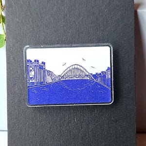 May include: A rectangular pin featuring a blue and white illustration of a bridge over water. The pin is set against a dark gray background, with a plant in the background. The bridge has an arched design, and the water is a solid blue color.