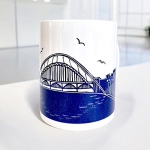 May include: White ceramic mug with a blue and white illustration of a bridge and cityscape. The mug has a handle on the right side.