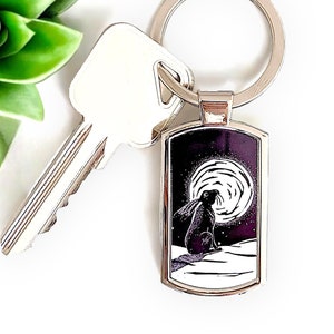 May include: Silver-toned keychain with a rectangular pendant featuring a black and white illustration of a hare under a full moon. The keychain is attached to a key and sits next to a green succulent.